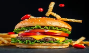 A close-up of a gourmet burger with a juicy beef patty, melted cheese, lettuce, tomato, and a sesame seed bun. Crinkle-cut fries and small red peppers are captured mid-air, falling onto the burger. The background is dark, highlighting the vibrant colors and textures of the food. The composition is dynamic and appetizing, emphasizing the fresh ingredients and dramatic presentation.