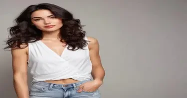 Create an image of a woman with long, dark, wavy hair, standing in a confident pose with one hand on her hip and her body slightly turned to the side. She has a striking face with almond-shaped, deep brown eyes, well-defined eyebrows, high cheekbones, and full lips with a natural, subtle smile. She is wearing a stylish, sleeveless white dress with a high slit on one side, revealing her toned leg clearly through the slit, and the dress features a delicate lace pattern with a tied detail at the chest. The background includes lush green plants and a modern outdoor setting, with soft natural lighting. Emphasize a natural, elegant look with a focus on her detailed facial features, the visibility of her leg through the dress slit, and the overall pose.