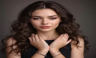 A young woman with long wavy brunette hair rests her head on her clasped hands. She wears a black top and a delicate bracelet on her left wrist. Her expression is soft and contemplative, with natural makeup and subtle pink lipstick. The background is dark and blurred, focusing on her face and hands