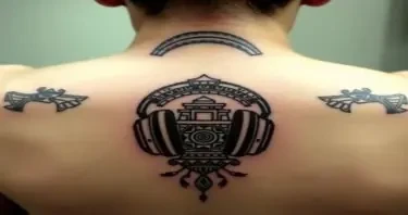 Anchored at the upper spine/base of the neck 	•	Curves outward across the trapezius to the tops of both shoulders 	•	Earcup designs sit just behind the shoulders — not on the scapulae 	•	There’s no tattoo on the back of the neck itself 	•	The central arch rests between C6–T1 vertebrae — right at the nape, but lower than typical collar ink 1. HEADPHONE TATTOO (NECK/UPPER BACk and chest) 	•	Style: Mayan-stylized headphones — thick curved arcs mimicking audio coils, wrapped in stepped temple glyphs.