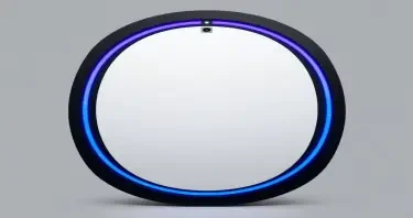 A tall, freestanding oval mirror unit measuring approximately 7 feet high and 3 feet wide, with a flawless, reflective surface. The mirror is framed in polished black onyx, matte in finish, with no visible seams. Two concentric LED-style rings encircle the mirror: the outer ring is electric blue and positioned just inside the frame, while the inner ring is violet and approximately 60% the diameter of the mirror. Between these rings, faint luminous markings and symbols suggest encoded information, softly pulsing. The top center of the unit houses a subtle projection module — a small inset lens that emits a faint vertical glow upward. The background is evenly lit in neutral gray to highlight the mirror without shadow. No human figure is present. The mirror stands in isolation as a sacred tech object.