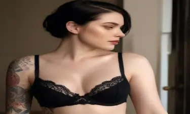 n a softly lit room, a striking young woman stands confidently, drawing attention with her intricate tattoos that elegantly adorn her neck and arms, contrasting beautifully against her pale skin. The deep black lace of her bra highlights her curves, while she poses with a subtle gaze, embodying a blend of allure and strength. Her dark hair cascades over her shoulders, framing her face which features smooth, radiant skin and an expressive aura. The warm tones of the room, coupled with the soft texture of the bedcover in the background, create an intimate atmosphere that enhances the allure of the scene, allowing the viewer to feel a captivating sense of mystery and charm.