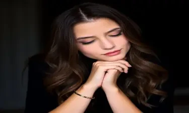 A young woman with long wavy brunette hair rests her head on her clasped hands. She wears a black top and a delicate bracelet on her left wrist. Her expression is soft and contemplative, with natural makeup and subtle pink lipstick. The background is dark and blurred, focusing on her face and hands