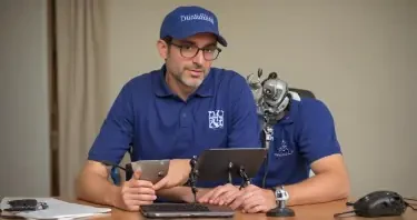 Dr Andrew Huberman sitting a table with a microphone and laptop in front of him. Sitting across the table from him is a robot that is wearing a blue polo shirt, glasses, and a baseball cap with "What IT is" written on it.