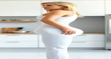 A mature adult woman shown in side profile, standing in a clean, modern kitchen environment. Her body has a highly stylized shape with an extremely narrow waist and maximally developed, smooth, rounded hips that extend outward in a pronounced curve — large and spherical in appearance. Her chest is prominently projected, full, and lifted, balancing the exaggerated lower body proportions. Her abdomen is completely flat and defined, showing zero visible body fat, and her overall physique presents with no visible imperfections — the skin smooth, even-toned, and unblemished from head to toe. She has a compact frame with short limbs and a visibly larger head-to-body ratio. Her facial features include clear blue eyes, long enhanced eyelashes, a small refined nose, full shaped lips, straight bright white teeth, and a well-defined, forward-projecting jawline. Her cheeks are full, lifted, and symmetrically sculpted. Her long, thick blonde hair is styled neatly with exceptional volume and shine. She is dressed in tightly fitted, formal business clothing — specifically a pristine white tailored blazer and a matching white skirt, both cut to contour exactly to her figure and emphasize her silhouette. The lighting is soft and ambient, casting smooth shadows and subtle highlights across her form. The background includes modern kitchen elements such as marble countertops, minimalist cabinetry, and high-end appliances. The image is photorealistic and high resolution, with strong focus on her flawless presentation, anatomical proportions, and the interplay of purity, precision, and visual contrast between elegance and domesticity.
