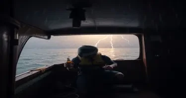 Man taking shelter inside the lower cabin of a docked old houseboat, crouched wearing a motorcycle helmet, life vest and clutching a coors light beer during a severe thunderstorm with torrential rain and strong winds