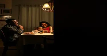 A split-image concept of a black family: On the left, a family laughing at a dinner table. On the right, a dimly lit, empty chair at the same table (symbolizing loss).