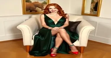 Redhead in dark green high slit plunging neck line gown  red high heel shoes and lace topped stockings held up by a matching green garter belt.  She is sitting in an arm chair dress pulled up to show her stockings. She smiles and wears a silver necklace and silver bracelets.