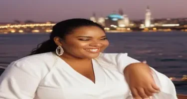A joyful SSBBW woman with a radiant smile, wearing a flowing white outfit with gold earrings, dining at a sunlit rooftop restaurant in Istanbul. She has braided hair, dark skin, and a confident, relaxed pose. The Bosphorus sparkles in the distance, and the city skyline glows in the warm evening light.